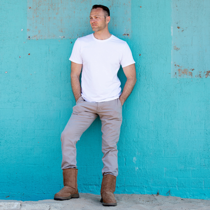 Man wearing a white t-shirt, beige pants, and brown boots against a turquoise wall.